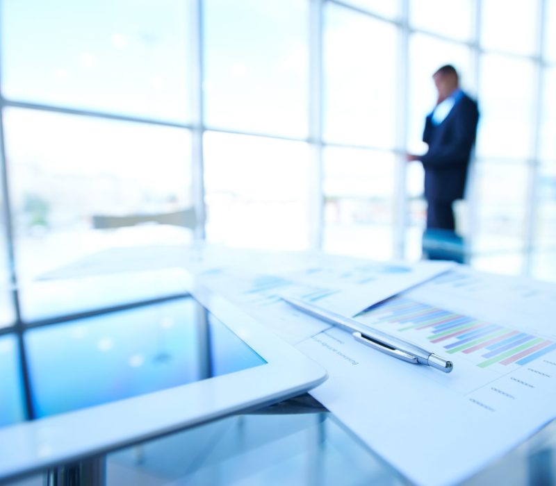 Business documents, touchpad and pen at workplace on background of businessman calling by the window